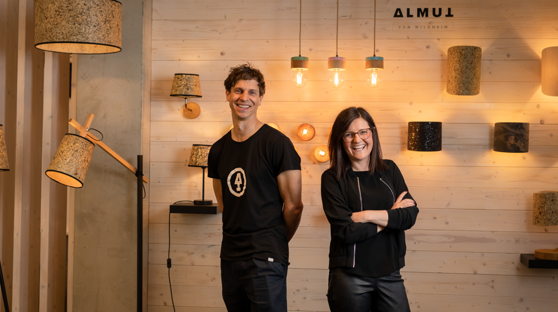 Steffi Graber & Ulrich Huber, stehen vor einer Wand mit Ihren Lampen und sehen lächelnd in die Kamera