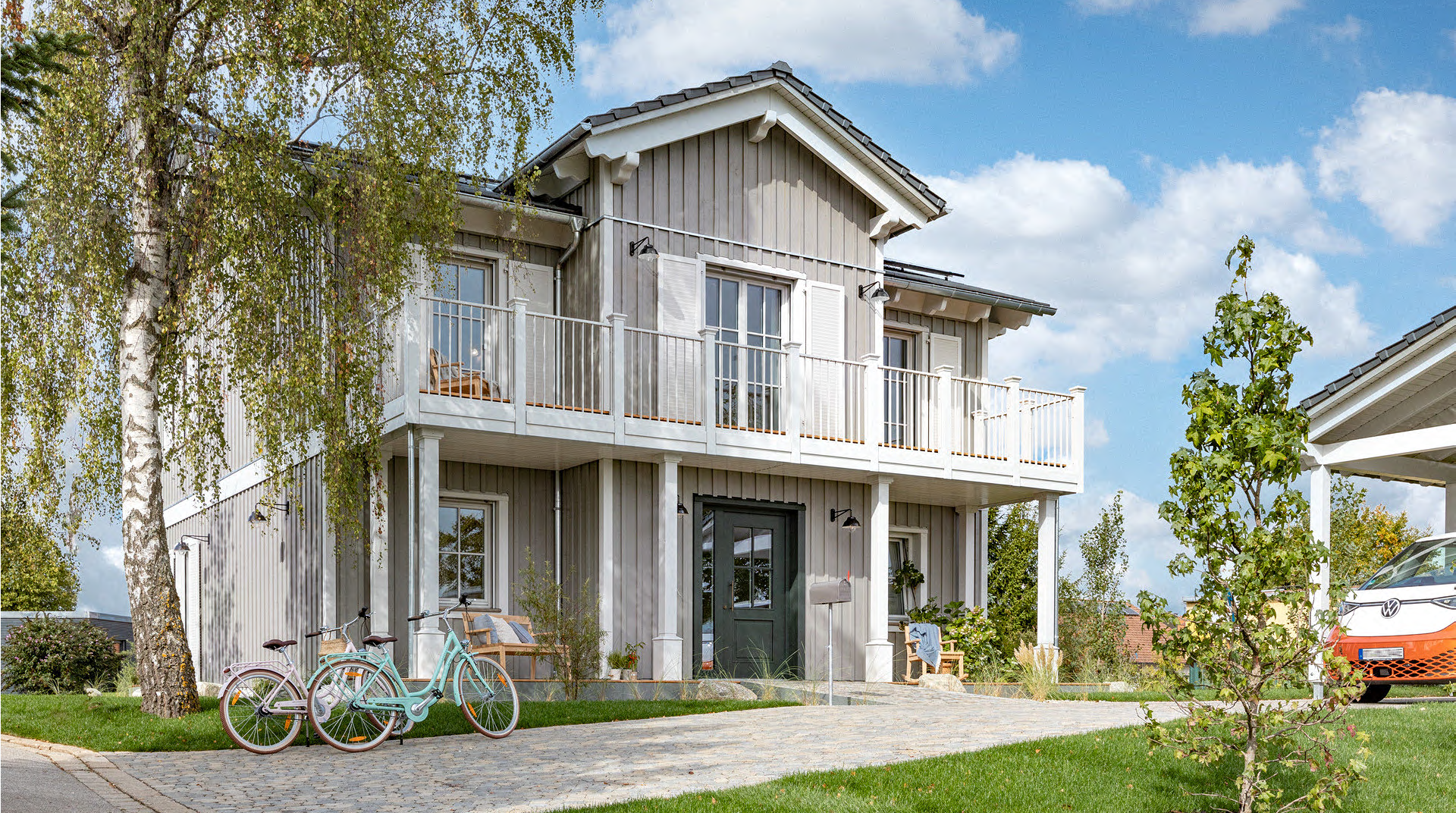 Frontansicht des Hauses. Die komplette Vorderseite ist überdacht durch einen Balkon, die Fassade ist aus Holz. Rechts sieht man den Carport. 