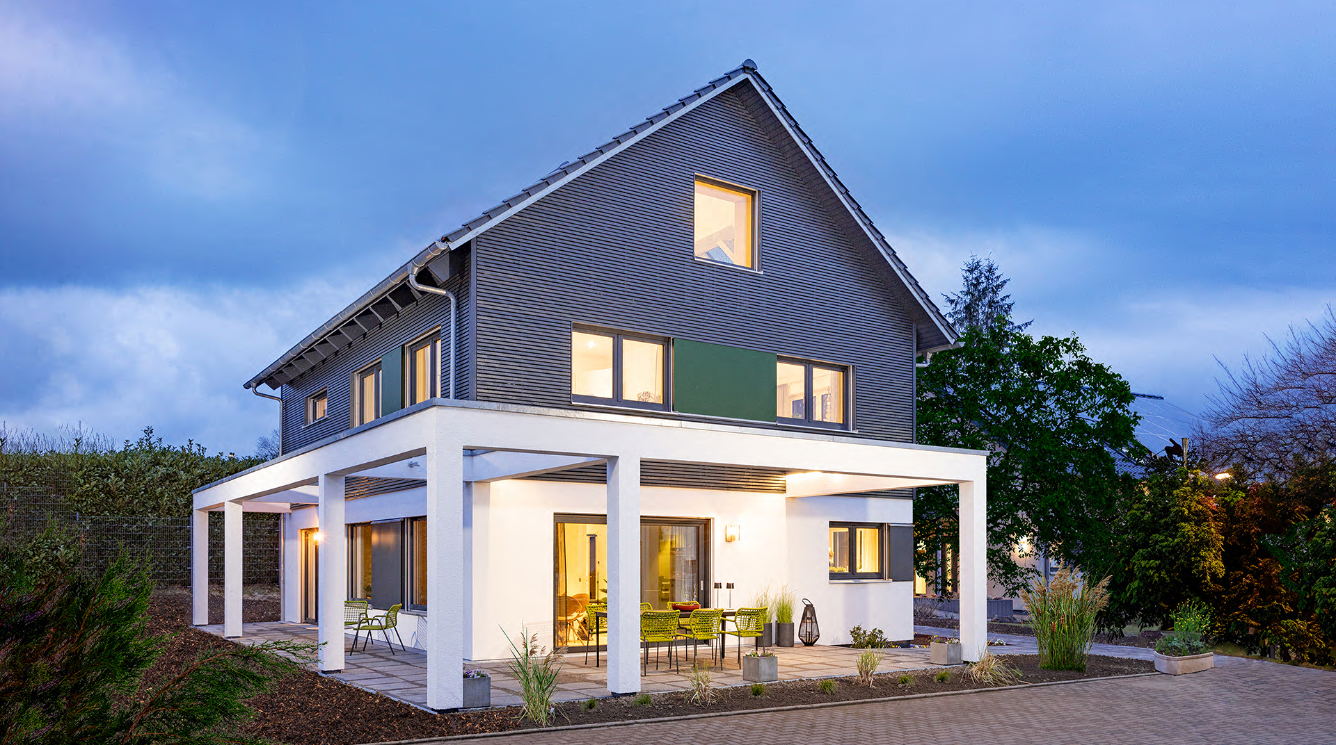 Das Haus abends von außen. Die große Terrasse um das Haus ist beleuchtet mit Tisch und Stühlen, umzogen vom Garten. Die untere Hälfte des Hauses ist hell Verputzt, oben ist es mit Holzlamellen verkleidet. Man sieht das Licht durch die Fenster scheinen.