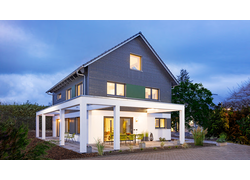 Das Haus abends von außen. Die große Terrasse um das Haus ist beleuchtet mit Tisch und Stühlen, umzogen vom Garten. Die untere Hälfte des Hauses ist hell Verputzt, oben ist es mit Holzlamellen verkleidet. Man sieht das Licht durch die Fenster scheinen.