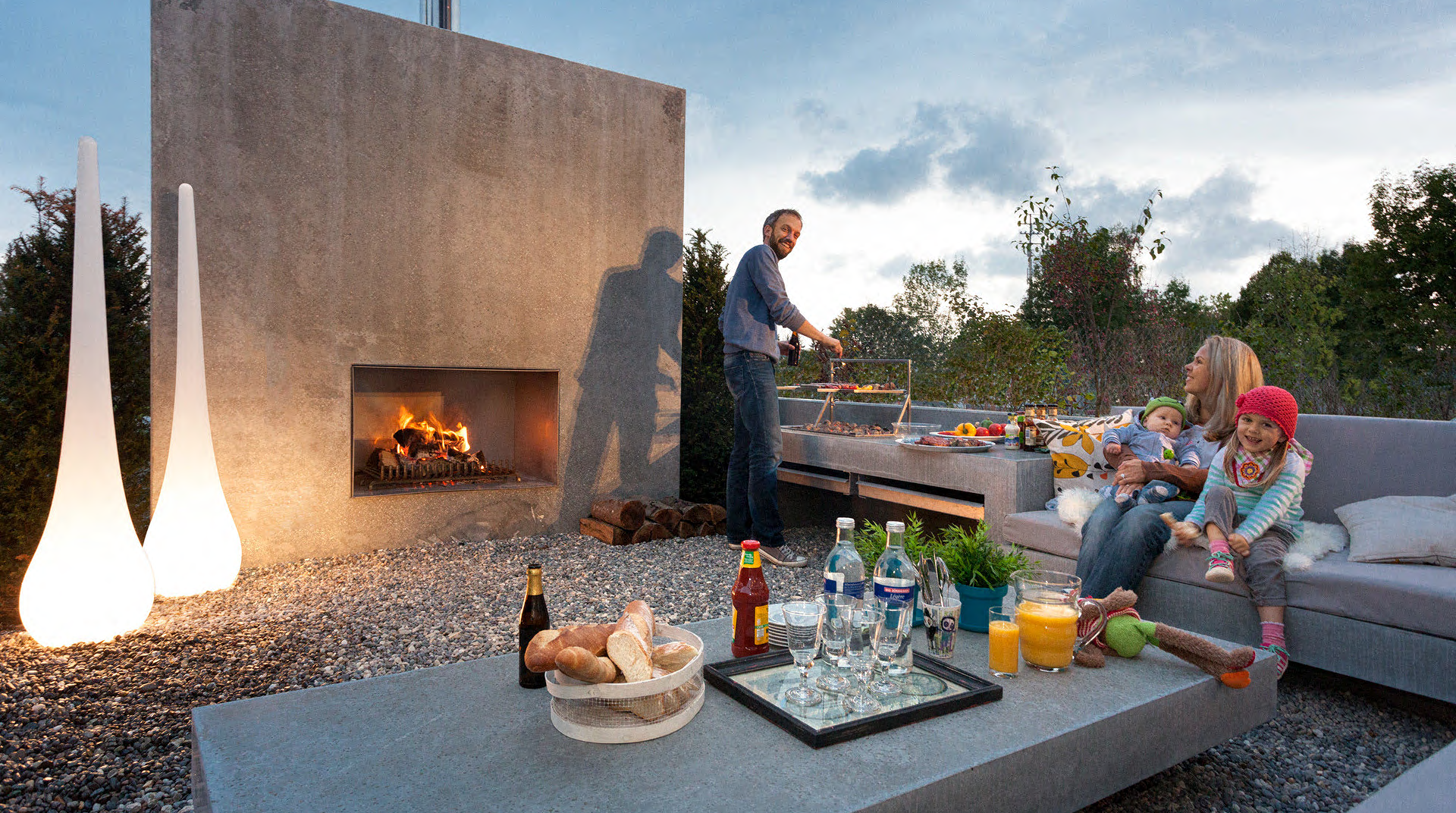 Familie auf der Terrasse fröhlich am Grillen mit Kaminfeuer im Hintergrund, einem großen Grill, Sofa und Tisch.